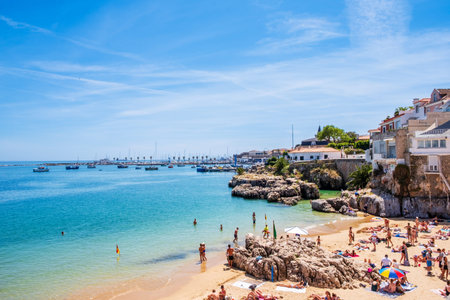 Cascais, Portugal - MAI 7, 2024: Tourists on sandy beach Praia da Rainha. Cascais harbor with sailboats in background, rocky coastline and vibrant coastal town under blue skyのeditorial素材