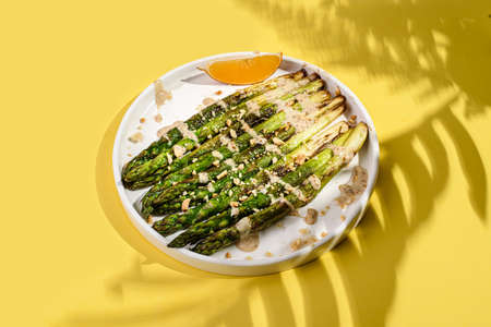 cooked asparagus in a plate. asparagus fried with olive oil, drizzled with sauce and sprinkled with nuts, next to a slice of lemon. On a yellow background, with shadows from branchesの写真素材