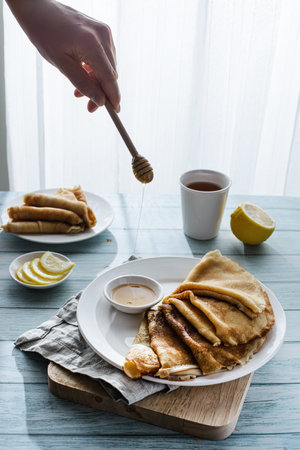 pancakes with honey on a wooden background. TEA , A WHOLE LEMON AND LEMON SLICES ARE ON THE TABLE Russian holiday maslenitsa, hand holds a honey spoonの写真素材