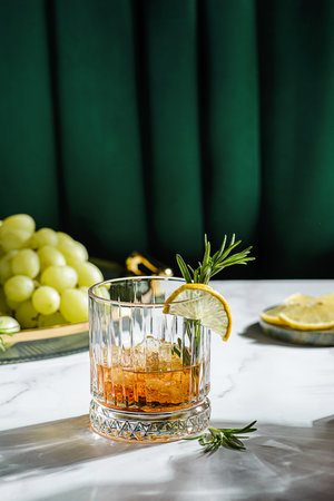 a glass of whiskey on the table is decorated with lemon and rosemary, There are grapes on a tray nearbyの写真素材