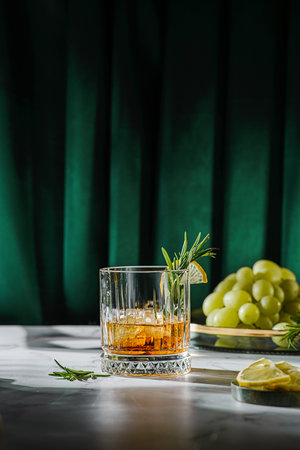 a glass of whiskey on the table is decorated with lemon and rosemary, There are grapes on a tray nearbyの写真素材