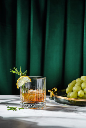 a glass of whiskey on the table is decorated with lemon and rosemary, There are grapes on a tray nearbyの写真素材