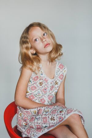 sad cute European teen girl on a gray background sitting on a red chair. beautiful curly blonde with white teeth.の写真素材