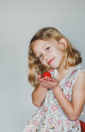 berry in the hands of a child. European blonde girl eating strawberries on a gray background. white teeth.の写真素材
