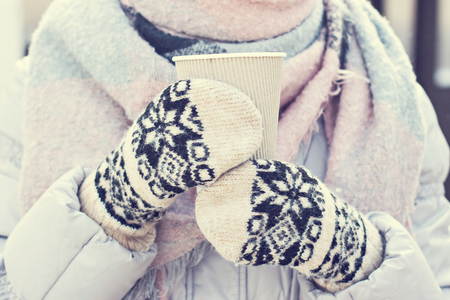 Female hands in white and black mittens holding steaming  cup of hot coffee cocoa, mulled wine or tea, outdoor. Winter time concept. Bask in the cold. Women's hands in mittens hold a craft paper glassの写真素材