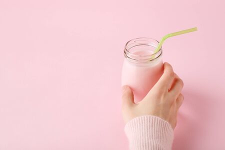 Women's hand holding glass bottle of fruit yogurt with straw on pastel pink background with copy space. Healthy drink concept, minimal.の写真素材