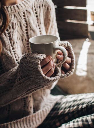 Cozy autumn or winter atmosphere. Young woman with cup of tea and warm sweater is seating at home near the window.の写真素材