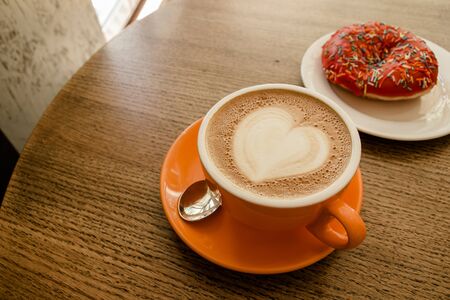 Cappuccino coffee with heart latte art and pink donut on wood table in cafe atmosphere.の写真素材