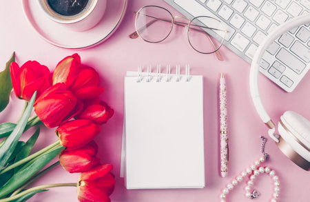 Home office workplace: cup of coffee, empty notebook, keyboard, tulip flowers, brushes, eyeglasses, beads, headphones on pink background. Flat lay, top view, copy spaceの写真素材