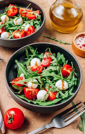 Healthy arugula salad with mozzarella cheese and cherry tomatoes in black bowls on beige rustic background. Selective focusの写真素材