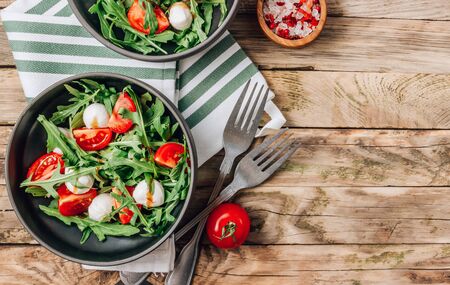 Healthy arugula salad with mozzarella cheese and cherry tomatoes in black bowls on wooden rustic background. Top viewの写真素材