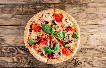 Sliced italian pizza with salami, mozzarella, mushroom, tomatoes, black olives and basil leaves on rustic wooden background. Italian traditional food. Popular street food concept. Top view. Minimalの写真素材