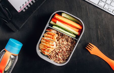 Healthy meal prep containers with quinoa, chicken and and sticks of cucumber and carrots on black wooden background. Healthy lunch at office. Take away concept. Top view.の写真素材