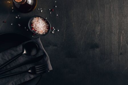 Black table setting: plate, napkin, silverware, olive oil and salt on black woodenの写真素材