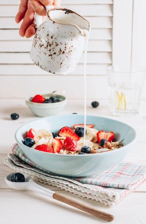 Milk is pouring into healthy breakfast bowl with oat granola with dried buckwheat, nuts and coconut chips, berries and rice milk on a white background. Healthy vegetarian eating. Selective focusの写真素材