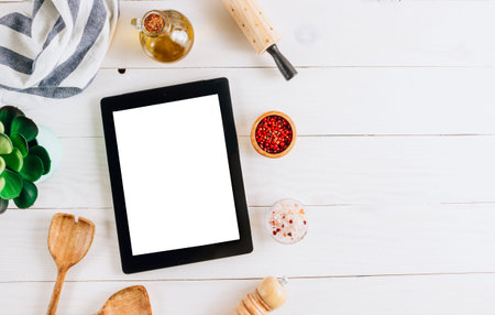 Tablet with blank screen and different kitchen and cooking utensils on whie wooden table. Culinary...