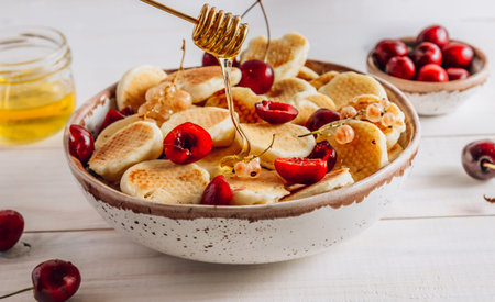 Trendy home breakfast with tiny pancakes in a ceramic bowl with honey and cherries on a white wooden...