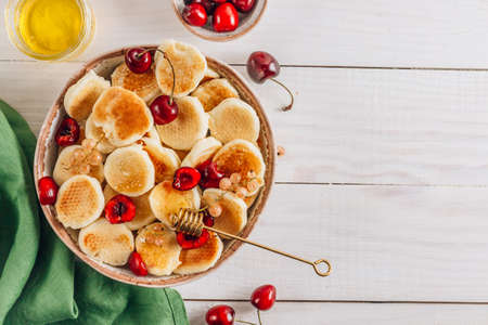 Trendy home breakfast with tiny pancakes in a bowl with honey and cherries on a white wooden table with a green napkin. Top view with copy spaceの写真素材