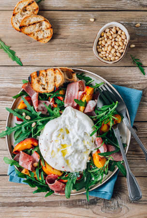 Heathy summer salad with grilled peach, arugula, ham and burrata on rustic wooden background. Summer food concept. top view.の写真素材