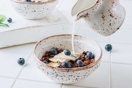 Chocolate granola and milk with blueberries, banana and mint over white tile background. Healthy breakfast concept. selective focusの写真素材