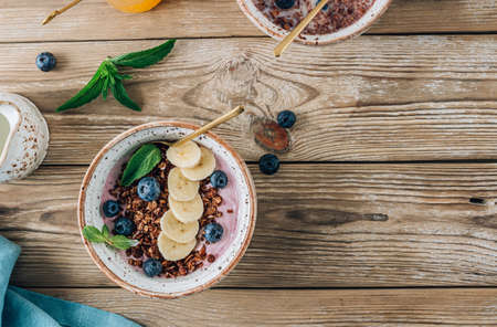 Berry banana smoothie bowl with chocolate granola, blueberries and mint over wooden background. Healthy breakfast concept. Top view with copy space.の写真素材