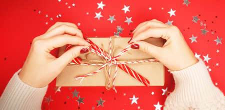 A Christmas gift in craft paper decorated with a Christmas cane in women's hands with a beautiful bright manicure, on a red background with scattered holiday stars.の写真素材