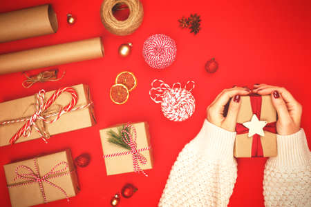 Christmas gift in kraft paper, decorated with a wooden star in womens hands with a beautiful bright manicure, on a red background with accessories for packaging.の写真素材
