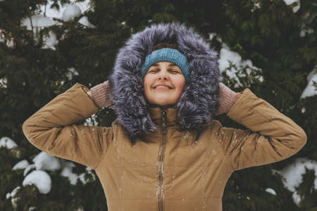 A smiling young woman in a knitted blue cap and a warm brown jacket looks up at the sky and enjoys the winter weather against the background of coniferous trees.の写真素材