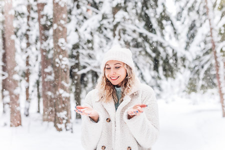 A young girl with a beautiful smile in white clothes catches snowflakes in a snowy forest in the...