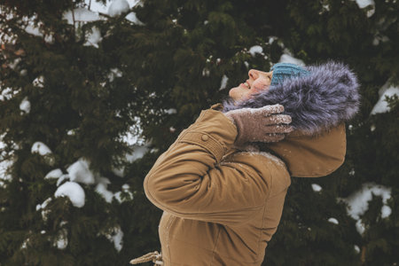 A smiling young woman in a knitted blue cap and a warm brown jacket looks up at the sky. She enjoys the winter weather against the background of coniferous trees.の写真素材