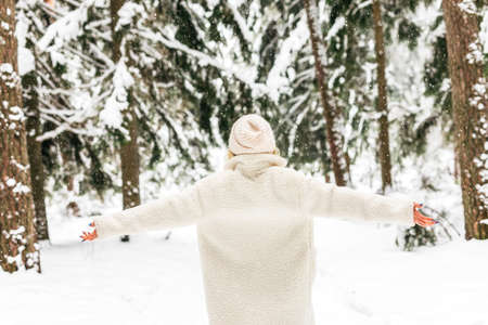 A young girl in a white hat and an artificial fur coat enjoys a walk through a snowy forest in cold winterの写真素材
