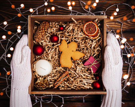 A girl in knitted white gloves holds a Christmas decorative gift in a wooden box with Christmas decorations and cookies in the shape of an angel.の写真素材