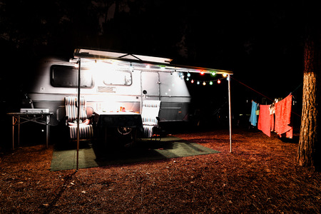 Retro vintage camping caravan trailer at night in a forest with lights, chairs and a tableの写真素材
