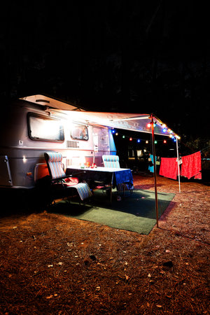 Retro vintage camping caravan trailer at night in a forest with lights, chairs and a table