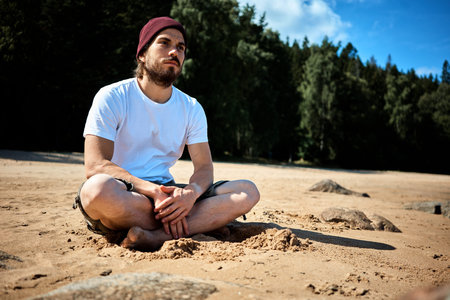 Young bearded man with hut and white shirt sitting at the beach in sweden and is looking thoughtful into the distance.の写真素材