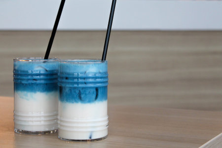 Two glasses of iced pea latte. Blue tea layered cocktail with milk on wooden table with black straw...