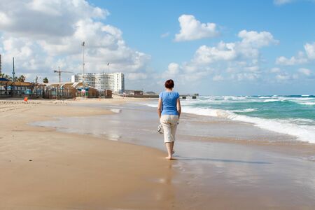 Bat Yam, Israel - 14 July, 2014. Slim old lady walking on the sandy beach. Mediterranean coastline. Travellers going barefoot through seafoam. Building reflected on the sand. Blue cloudy sky.のeditorial素材