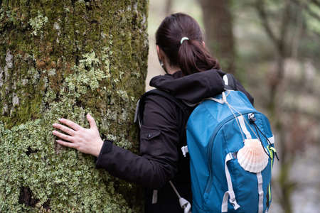 White woman with ponytail, doing the Camino de Santiago, dressed in a black jacket, with a blue backpack, with a hanging shell, symbol of the pilgrim, hugging a treeの写真素材