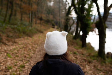 Brunette woman, walking through a forest, on a path full of leaves on the ground, wearing a black jacket and a white hat shaped like a cat. Lifestyle conceptの写真素材