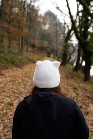 Brunette woman, walking through a forest, on a path full of leaves on the ground, wearing a black jacket and a white hat shaped like a cat. Lifestyle conceptの写真素材