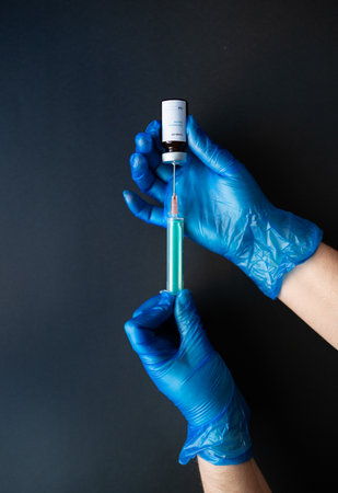 A nurse wearing blue gloves, holding a vial of coronavirus vaccine with one hand, and with the other a needle, with a black backgroundの写真素材