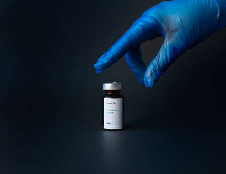 A hand of a nurse wearing a blue glove, picking up a vial of coronavirus vaccine, with a black backgroundの写真素材