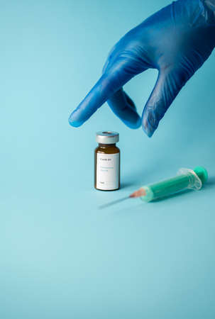 A hand of a nurse wearing a blue glove, picking up a vial of coronavirus vaccine, a syringe with a needle, with a blue backgroundの写真素材