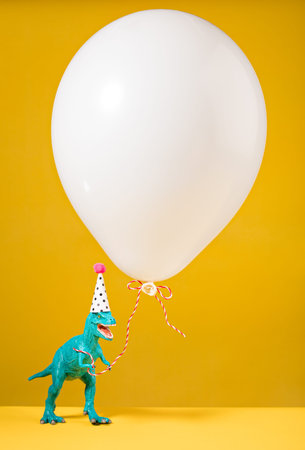Teal dinosaur toy with birthday hat holding a white balloon on a yellow background.の写真素材