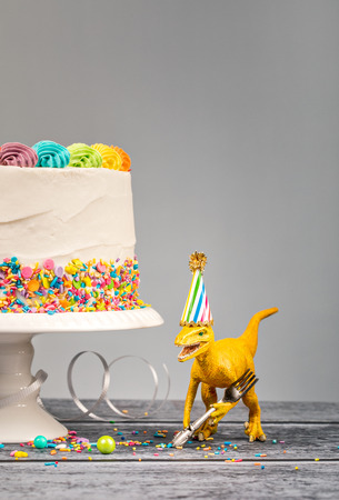 Hungry toy dinosaur wearing a hat and holding a fork next to a birthday Cake on a grayの写真素材