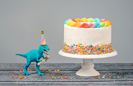 Hungry toy dinosaur wearing a hat and holding a fork next to a birthday Cake on a grayの写真素材