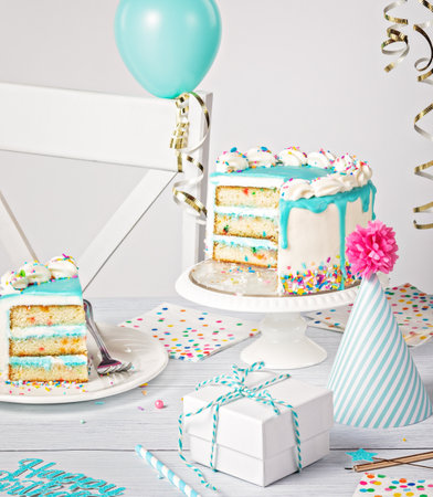 Birthday party table setting with a teal blue ganache drip cake on a light grey white background with sliced piece of cake and decorations.の写真素材