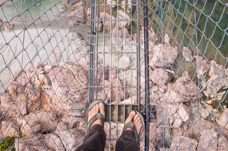 Looking down on feet in flip-flops on a high and dangerous airwalk with a river and rocks belowの写真素材