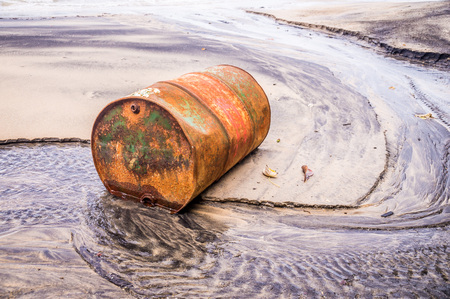 Rusty barrel oil on a partly black coloured beach illustrates the pollution of environment by oil spillsの写真素材