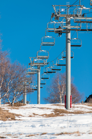 A man takes a ride in a ski lift up the hill on a day in spring with few snow and blue skyの写真素材